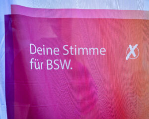 Das Bild zeigt den Ausschnitt einer Flagge des BSW weiß mit einem Verlauf von Pink zu Orange und der Aufschrift Deine Stimme für BSW.