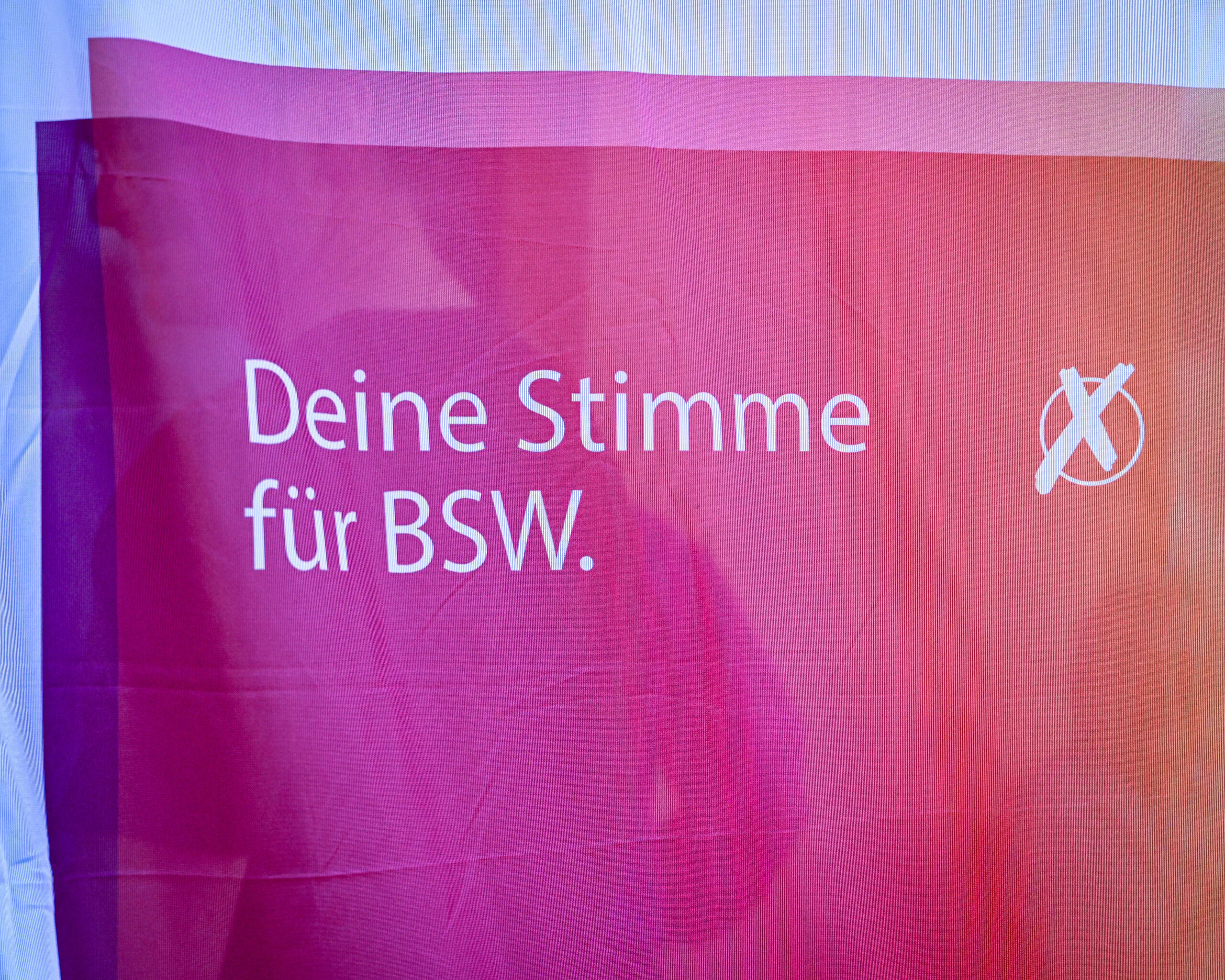 Das Bild zeigt den Ausschnitt einer Flagge des BSW weiß mit einem Verlauf von Pink zu Orange und der Aufschrift Deine Stimme für BSW.