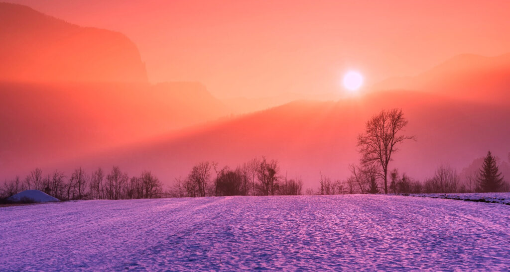 Sonnenuntergang in sanftem Dunst mit Hügeln über einem Schneefeld, Farben von Orange am Himmel bis Violett auf dem Schneefeld. Am Ende des Feldes ist eine dunkle Baumreihe von kahlen Laubbäumen zu sehen.